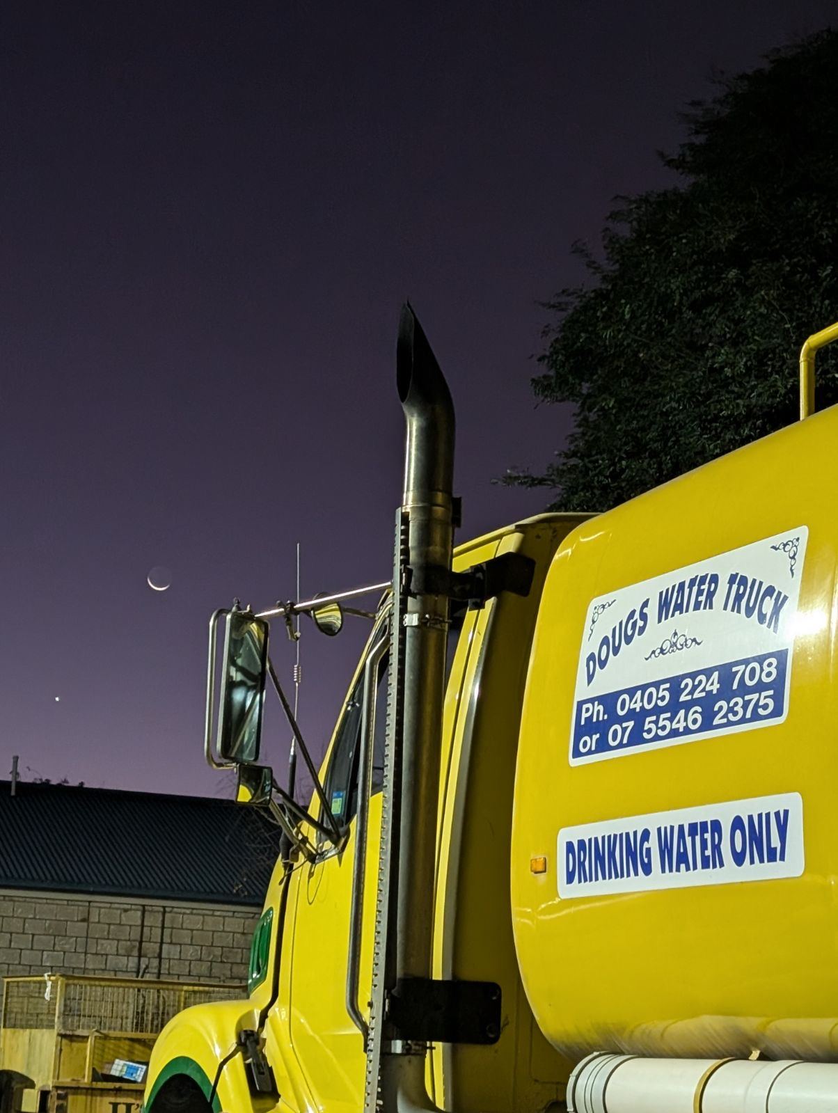Yellow Dougs Water Truck cab and exhaust stack at dusk beneath a crescent moon